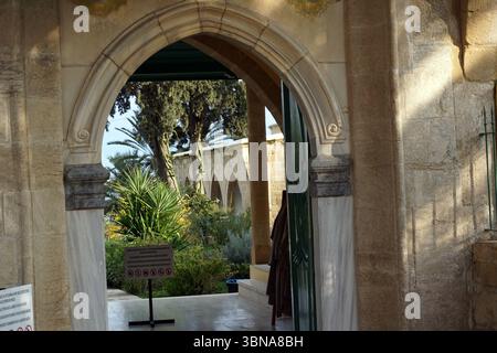 Hala Sultan Tekke. Une arche en pierre, probablement partie d'un bâtiment, avec une porte verte et un panneau indiquant une zone restreinte. L'arche est encadrée par un mur de pierre, et une partie d'un bâtiment avec des fenêtres cintrées est visible à travers l'arche. L'arche est entourée d'une variété de plantes et d'arbres, y compris des palmiers, visibles en arrière-plan. Un panneau avec un cercle rouge et une barre oblique est placé sur un stand à proximité. Le sol semble pavé de pierre ou de béton. Légende façonnée par l'œil d'un artiste et l'imagination., Chypre, lac salé de Larnaca Banque D'Images