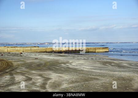 Chypre, Larnaca, Une scène de plage sereine. La plage de sable, avec sa teinte brun clair, s'étend vers l'océan bleu clair et calme. Une digue en béton, construite de grands blocs, se dresse au premier plan, avec quelques petits rochers éparpillés autour. L’océan est relativement calme avec de petites vagues qui se battent doucement contre le rivage. Au loin, quelques bateaux peuvent être vus sur l'eau. Le ciel est d'un bleu clair avec quelques nuages dispersés, et l'éclairage général suggère que c'est probable Banque D'Images