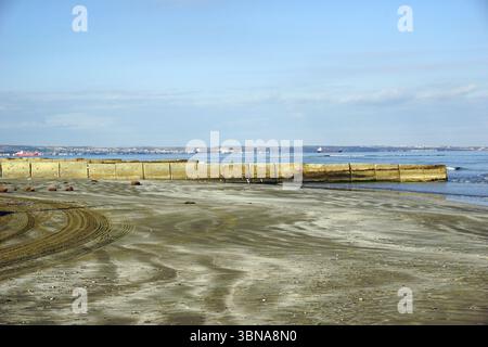 Chypre, Larnaca, Une scène de plage sereine. La plage de sable, avec sa teinte brun clair, s'étend vers l'eau calme et bleu clair. Une digue en béton, construite de grands blocs, se dresse au premier plan, avec quelques petits rochers éparpillés autour. L'eau est relativement calme, avec de douces vagues qui rodent contre le rivage. Au loin, quelques bateaux peuvent être vus sur l'eau. Le ciel est d'un bleu clair avec quelques nuages dispersés, et l'horizon est visible dans le Banque D'Images
