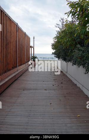 Chypre, Larnaca, Une scène sereine en bord de mer. Une passerelle en bois, faite de planches, s’étend vers l’océan, flanquée d’une clôture en bois à gauche et d’un mur blanc à droite. Une petite plante en pot est placée sur la passerelle près de la clôture. L'océan est visible en arrière-plan, avec une plage de sable et un ciel nuageux au-dessus. La passerelle est bordée par des buissons et des arbres verdoyants, créant une atmosphère tranquille. Un œil d'artiste et une imagination façonnée Banque D'Images