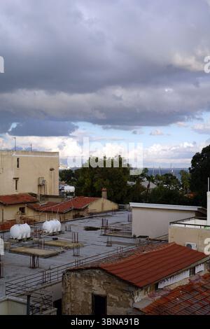 Un paysage urbain avec un mélange de bâtiments anciens et neufs. Le ciel est rempli de nuages, projetant une lumière douce sur la scène. Les bâtiments sont principalement de couleur beige ou crème, certains présentant des toits de tuiles rouges. Plusieurs bâtiments ont des toits plats, et certains ont des cheminées. Au loin, la mer est visible, avec quelques voiliers ou yachts à l'horizon. Les bâtiments sont situés sur un toit, certaines structures ayant des ossatures métalliques exposées et d'autres avec des matériaux de toiture plus traditionnels. L'image est prise d'un point de vue élevé, offrant une vue complète du paysage urbain et de son arc Banque D'Images
