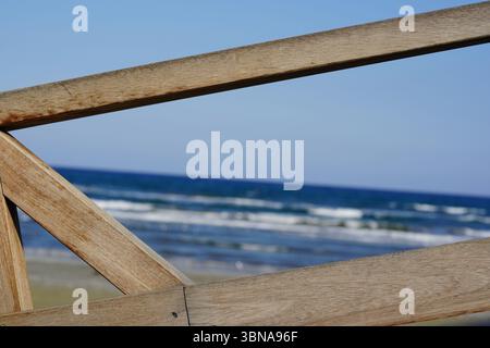 Chypre, Larnaca, Une balustrade en bois, peut-être partie d'un stand de sauveteur, avec une forme triangulaire et un aspect légèrement altéré. La balustrade est positionnée au premier plan, avec la vaste étendue de l'océan et un ciel bleu clair formant la toile de fond. L'océan est d'un bleu profond, avec des vagues blanches qui rodent doucement contre le rivage. La ligne d'horizon est clairement visible, séparant l'océan du ciel. La perspective de l'image est à partir d'un angle bas, regardant vers le haut de la rampe, ce qui améliore la sensation de hauteur et de grandeur de la scène. Le sable sur la plage est de couleur beige clair ou beige. An Banque D'Images