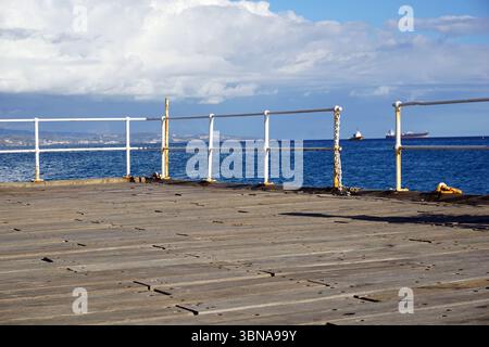 Limassol, Chypre, Une jetée en bois s'étendant dans une vaste étendue d'océan bleu. La jetée est construite à partir de planches de bois et comporte une rampe en métal blanc et des poteaux en métal peint en jaune. Le garde-corps est soutenu par un poteau en métal rouillé. Le ciel ci-dessus est d'un bleu clair, parsemé de nuages blancs moelleux. Au loin, deux navires peuvent être vus à l'horizon. La perspective de l'image est de la jetée, regardant vers le Banque D'Images