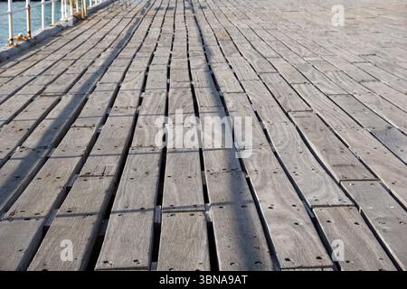 Limassol, Chypre, Une vue rapprochée d'une jetée en bois s'étendant au loin. La jetée est construite à partir de planches de bois gris-brun altérées disposées en rangées parallèles, créant un motif rythmique. Les planches sont légèrement décalées les unes par rapport aux autres, ajoutant une sensation de profondeur à l'image. La perspective est du sol, regardant vers le haut la jetée, qui semble être faite d'un matériau composite. Les planches montrent des signes d'altération et d'usure, certaines zones apparaissant plus foncées ou plus claires que d'autres. En arrière-plan, un plan d'eau est visible, avec une partie d'une rampe ou d'une clôture le long Banque D'Images