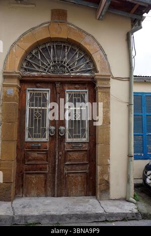 Une porte en bois vieilli avec une grille décorative en métal, sertie dans une arche en pierre. La porte est flanquée de deux piliers en pierre robustes, et une petite plaque est visible sur le pilier gauche. La porte a deux ensembles de portes doubles, chacun avec une grille décorative en métal. L'arche en pierre au-dessus de la porte est ornée d'une grille décorative en métal. La porte est placée dans un mur beige clair ou crème. Une partie d'une fenêtre bleue avec des volets blancs est visible à droite de la porte. Une petite section d’une voiture de couleur foncée est garée à droite de la porte., Nicosie (Lefkosia), Chypre, la dernière division du monde Banque D'Images