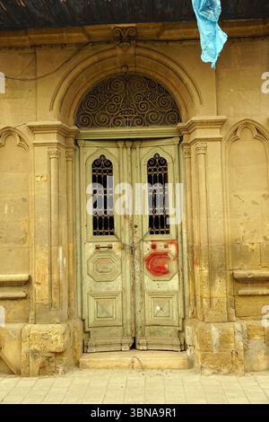 Nicosie (Lefkosia), Chypre, la dernière capitale divisée du monde, porte en bois vert clair altérée surmontée d’une grille décorative en métal, située dans une arche en pierre. La porte est légèrement entrouverte, révélant un aperçu de l'intérieur. La voûte en pierre est flanquée de deux colonnes de pierre, et une bâche bleue est suspendue au plafond au-dessus. La porte est légèrement entrouverte, et une petite tache rouge est visible sur la porte droite. La voûte en pierre fait partie d'une structure en pierre plus grande, et le sol devant la porte est fait de pierre de couleur claire ou Banque D'Images