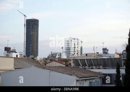 Nicosie (Lefkosia), Chypre, la dernière capitale divisée du monde, Une ligne d’horizon au crépuscule, avec un grand bâtiment cylindrique en construction, sa forme squelettique se démarquant des tons doux du ciel. Le bâtiment est partiellement recouvert d'échafaudages, indiquant des travaux en cours. En arrière-plan, un mélange de bâtiments avec différents styles architecturaux et couleurs, y compris un bâtiment blanc avec une façade courbée, sont visibles. Le ciel est d'un bleu clair avec quelques nuages dispersés. Le premier plan est dominé par une rangée de bâtiments aux toits en terre cuite, et quelques arbres sont visibles dans le Banque D'Images