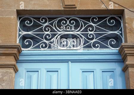 Nicosie (Lefkosia), Chypre, dernière capitale divisée du monde, porte en bois bleu clair surmontée d’une grille décorative en métal blanc. La grille présente un design en forme de coeur avec des motifs tourbillonnants et le numéro '1908' inscrit à l'intérieur. La porte est placée dans un mur de pierre beige, avec une partie d'une colonne de pierre brune visible sur le côté gauche. La porte est fermée et l'image est prise sous un angle légèrement bas, en regardant la porte et la grille décorative. Un œil d'artiste et une imagination façonnée Banque D'Images