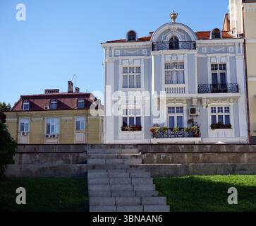 Plovdiv, la deuxième plus grande ville de Bulgarie, est l'une des plus anciennes villes d'Europe habitées en permanence, avec une histoire de plus de 8 000 ans. Niché le long de la rivière Maritsa., Un quartier résidentiel serein avec un bâtiment blanc proéminent avec un toit rouge et des détails ornés, y compris un finial décoratif sur le toit. Le bâtiment dispose d'un balcon au deuxième étage avec une balustrade en fer forgé noir et des jardinières. Il est sur une zone herbeuse avec un mur de soutènement en pierre et un ensemble d'escaliers menant à elle. A gauche, un bâtiment beige avec un toit rouge et un petit balcon est visible. A droite, un jaune Banque D'Images