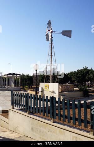 Une scène de parc sereine. Au premier plan, un grand moulin à vent blanc avec un axe noir et une lame argentée se tient sur une plate-forme en béton gris clair. Une clôture en bois vert foncé longe l'avant de la plate-forme, et une petite structure en pierre ou en béton de couleur claire est située derrière. Une lampe globe blanche est positionnée près du moulin à vent. L'arrière-plan présente un ciel bleu clair et des arbres verdoyants, créant une atmosphère tranquille. Une passerelle pavée est visible à mi-sol, et une partie d'un bâtiment ou d'une structure est visible au loin. Une légende en forme d'œil d'artiste et d'imagination., Paral Banque D'Images