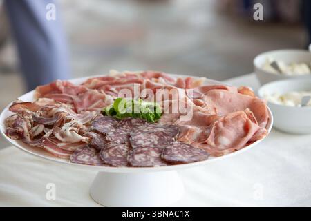 Tranches de salami, mortadelle, bacon et légumes verts frais magnifiquement disposés sur un plateau blanc, créant une alléchante présentation en hors-d'œuvre ou en-cas Banque D'Images