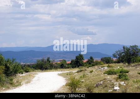 Un paysage serein avec un chemin de terre serpentant à travers un champ herbeux parsemé de rochers. Le chemin mène à une colonie nichée dans la vallée, avec des bâtiments aux toits rouges. La colonie est entourée d'un mélange d'arbres et d'arbustes, et les montagnes en arrière-plan sont d'une couleur bleu-gris foncé. Le ciel est rempli de nuages, projetant une lumière douce sur la scène. Les montagnes au loin semblent recouvertes de végétation, et la palette de couleurs globale de l'image est dominée par des nuances de bleu, de gris et de vert. Un œil d'artiste et l'imagination façonné légende., Bansko est un pittoresque Banque D'Images