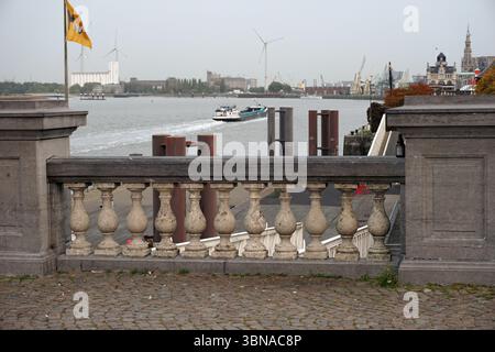 Une vue depuis une balustrade en pierre surplombant un port. La balustrade dispose d'une série de balustres ronds et d'une balustrade, offrant une sensation de profondeur et de perspective. Au-delà de la balustrade, le port est animé par l'activité, y compris un bateau blanc et bleu naviguant sur l'eau, et un ferry ou une barge rouge et blanc. Le port est situé sur une zone pavée ou pavée de briques, et l'arrière-plan comprend une ligne d'horizon avec des bâtiments et des éoliennes, suggérant un mélange d'éléments urbains et industriels. Un drapeau jaune avec du texte noir est visible sur un poteau près du côté gauche de l'image., Anvers, Secon Banque D'Images