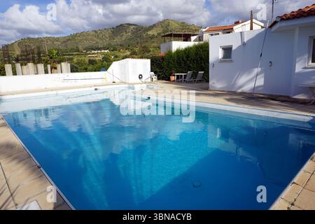 Argaka, Chypre, Une piscine rectangulaire avec une teinte bleu clair/turquoise, entourée d'un mur blanc, avec une petite table et deux chaises placées à proximité. Un bâtiment blanc avec un toit rouge est visible à l'arrière-plan, ainsi qu'une petite structure blanche, éventuellement de stockage ou utilitaire. Une clôture blanche ou un mur est également visible derrière la piscine. Le patio est bordé par un mur bas en pierre ou en béton de couleur claire. Au loin, une colline avec un mélange de végétation verte et quelques zones brunes/bronzées est visible. Le ciel est d'un bleu clair avec quelques blancs Banque D'Images