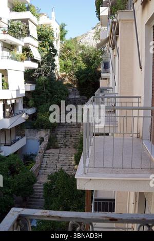 Un escalier étroit et sinueux qui monte à travers un paysage verdoyant et luxuriant. L'escalier est flanqué d'immeubles blancs avec balcons, et les bâtiments sont ornés d'auvents et de verdure. La perspective est d'un angle bas, regardant vers le haut l'escalier, qui est fait de pierre et a une balustrade en métal sur le côté droit. L'escalier est entouré d'arbres et de buissons, et un petit bâtiment de couleur claire est visible en arrière-plan. Le ciel est d'un bleu clair et clair. Un œil d'artiste et une légende façonnée par l'imagination., Athènes — la capitale vibrante de la Grèce, imprégnée d'histoire et de culture, A Banque D'Images