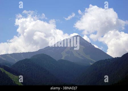 Bansko est une ville pittoresque dans le sud-ouest de la Bulgarie, nichée au pied des montagnes Pirin., Une chaîne de montagnes majestueuse sous un ciel bleu clair. La montagne, avec son sommet fortement pointu, domine la scène. Ses pentes sont couvertes d'arbres verdoyants, contrastant avec le ciel bleu parsemé de nuages blancs moelleux. Le sommet de la montagne semble avoir un terrain rocheux ou inégal. L'image est prise à distance, offrant une vue panoramique sur la chaîne de montagnes et son paysage environnant. Les montagnes sont principalement couvertes d'arbres vert foncé, avec certaines zones montrant des notes de Banque D'Images