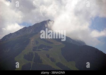 Bansko est une ville pittoresque dans le sud-ouest de la Bulgarie, nichée au pied des montagnes Pirin., Une chaîne de montagnes majestueuse avec un pic proéminent partiellement obscurci par les nuages. La montagne est couverte d'un mélange de végétation verte et brune, avec une station de ski visible sur ses pentes. La station de ski dispose d''une remontée mécanique et de plusieurs bâtiments, ce qui en fait une destination populaire pour les amateurs de sports d''hiver. Le ciel est d'un bleu clair avec des nuages blancs dispersés, et le sommet de la montagne est partiellement enveloppé dans une couche de nuages. L'image est prise à partir d'un point de vue élevé, fournissant une vue complète Banque D'Images