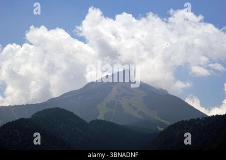 Bansko est une ville pittoresque dans le sud-ouest de la Bulgarie, nichée au pied des montagnes Pirin., Une chaîne de montagnes majestueuse sous un ciel bleu clair. La montagne, avec son pic rocheux accidenté, est partiellement obscurcie par une couche de nuages blancs. Les pentes de la montagne sont couvertes d'arbres verdoyants, et une remontée mécanique ou une structure similaire est visible sur la face de la montagne. La perspective de l'image est de loin, regardant la montagne, soulignant sa grandeur. Le ciel est d'un bleu clair et lumineux, et le sommet de la montagne est visible au centre de l'image. L'œil et l'imagination d'un artiste Banque D'Images