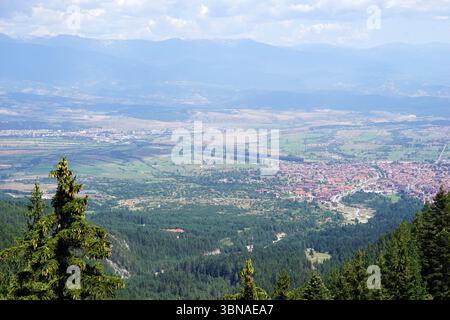 Une vue panoramique sur un paysage montagneux, avec un groupe de bâtiments nichés dans la vallée ci-dessous. Les bâtiments présentent un mélange de toits rouges et blancs, entourés de champs verdoyants et d'arbres. Une route ou un chemin sinueux est visible dans la zone. Les montagnes en arrière-plan sont un mélange de vert et de brun, avec quelques taches de ciel bleu visibles. Le point de vue est élevé, offrant une vue panoramique sur les bâtiments et le paysage environnant. Le ciel est bleu clair avec quelques nuages blancs. Un œil d'artiste et l'imagination façonné légende., Bansko est une ville pittoresque dans le sud-ouest de Bulgari Banque D'Images