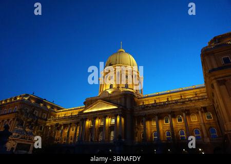 Statue équestre d'Eugène de Savoie qui se dresse en face de la Galerie nationale hongroise sur la colline du château de Budapest vue dans la soirée. Une grande structure avec un dôme doré et une série de colonnes, debout majestueusement contre un ciel bleu profond. Le bâtiment est éclairé par des lumières chaudes et dorées, créant un contraste saisissant avec l'obscurité environnante. La perspective de l'image est d'un angle bas, soulignant la stature imposante du bâtiment. La façade du bâtiment est ornée de nombreuses fenêtres, et une partie d'une statue ou d'un monument est visible sur le côté gauche de l'image., Buda Banque D'Images