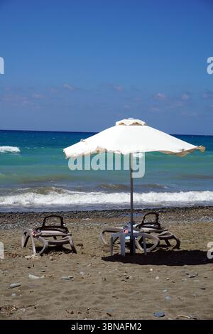Cyprus Beach, Une scène de plage sereine. Au premier plan, un parapluie blanc est ouvert, offrant de l'ombre sur une petite table avec une nappe blanche. Deux chaises longues vides sont placées sous le parasol. La plage est couverte de sable brun clair, avec de petits cailloux dispersés à travers elle. L'océan, d'une couleur bleu-vert profond, s'étend en arrière-plan, avec de petites vagues qui se balancent doucement contre le rivage. Le ciel est d'un bleu clair et lumineux, et l'horizon est visible dans le Banque D'Images