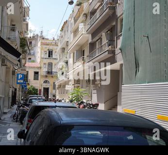 Une rue étroite dans un cadre urbain, bordée de bâtiments dans les tons beige, crème et jaune. Les bâtiments sont dotés de balcons et d'auvents, et certains possèdent des antennes paraboliques. Quelques voitures garées sont visibles le long de la rue, et quelques motos sont garées sur le trottoir. Un panneau de stationnement bleu est visible sur le côté gauche de la rue. La rue est pavée de pavés, et une partie d'un auvent vert ou d'une bâche est visible sur le côté droit de l'image. Le ciel est d'un bleu clair, et la scène est baignée de lumière naturelle. Légende façonnée par l'œil d'un artiste et l'imagination. Athènes, la capitale dynamique Banque D'Images