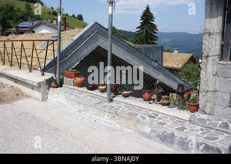 Une scène pittoresque et rustique avec une structure de toit en métal gris avec une section triangulaire. Le toit est partiellement couvert par une clôture à maillons de chaîne, et une petite structure de couleur sombre est visible derrière. Le toit est soutenu par un mur de béton avec une bordure en pierre. Un petit objet de couleur sombre, peut-être un nichoir, est attaché au mur. Le sol devant le toit est pavé de gravier ou de petites pierres. Plusieurs pots en terre cuite et un petit animal de couleur foncée, peut-être un cheval, sont placés sur le sol près de la clôture. Un petit objet de couleur foncée, peut-être une pelle, est également visible près du f Banque D'Images