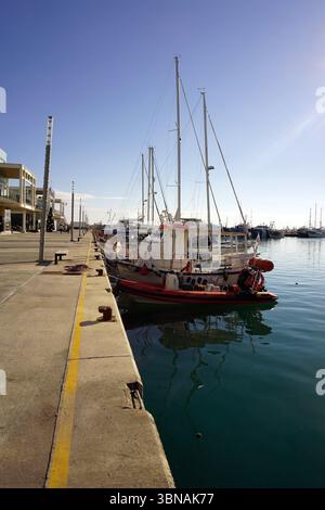 Limassol, Chypre, Une scène portuaire sereine avec une variété de bateaux amarrés le long d'une jetée en béton. La jetée est bordée par une ligne jaune et comporte quelques petits objets de couleur sombre, éventuellement des bouées ou des ancres. L'eau calme reflète le ciel bleu clair au-dessus, créant une atmosphère tranquille. Les bateaux, dont un petit bateau rouge et blanc, un voilier blanc avec une bande rouge, et un autre voilier blanc avec une bande rouge, sont amarrés le long de la jetée. En arrière-plan, quelques bâtiments sont visibles, et quelques arbres peuvent être vus le long du rivage. Le ciel est clair, bleu vif, et la scène globale Banque D'Images