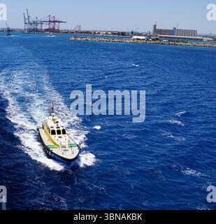Limassol, Chypre, Un bateau blanc et jaune avec une bande rouge et verte sur le côté, coupant à travers l'océan bleu profond. Le bateau se déplace vers le côté droit de l'image, laissant une traînée de mousse blanche dans son sillage. Au loin, un port avec plusieurs grues et un grand bâtiment est visible. Une petite île avec un rivage rocheux est également visible au milieu à droite de l'image. Le ciel est d'un bleu clair et vif. Un œil d'artiste et une imagination façonnée Banque D'Images