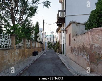Nicosie (Lefkosia), Chypre, la dernière capitale divisée du monde, Une rue étroite pavée dans un style rappelant l'architecture méditerranéenne. La rue est flanquée de bâtiments avec des murs blancs et quelques murs beiges ou beiges, créant un mélange harmonieux de couleurs. Sur le côté gauche, une clôture blanche avec un portail décoratif en métal est parallèle à la route. Sur le côté droit, un mur de pierre avec un panneau fané indiquant 'ANT 20' est visible. La rue est bordée d'arbres et de buissons, et quelques petites plantes sont visibles le long du mur. Les poteaux électriques et les lignes électriques sont également visibles le long de la rue. Le ciel est un Banque D'Images