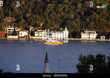 Istanbul, Turquie. La plus grande ville de Turquie et l'une des rares villes transcontinentales au monde, couvrant à la fois l'Europe et l'Asie à travers le détroit du Bosphore., Une scène sereine de front de mer avec un grand yacht orange et blanc naviguant sur l'eau bleue profonde. Le yacht est positionné près du centre de l'image, créant une sensation de profondeur. L'eau est relativement calme, avec seulement un petit sillage derrière le yacht. En arrière-plan, un groupe de maisons blanches et rouges aux toits rouges est niché à flanc de colline, entouré d'arbres verdoyants. Une église avec une flèche haute et pointue est visible au milieu du sol, et Banque D'Images