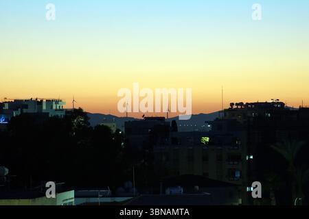 Larnaca, Chypre, l'image capture un paysage au coucher du soleil, avec le ciel peint dans des tons orange et bleu. C'est un ensemble de bâtiments, principalement des structures de faible hauteur, avec quelques bâtiments plus hauts visibles au loin. Plusieurs éoliennes sont visibles au milieu du sol, ajoutant un élément dynamique à la scène. La zone est surélevée, avec des montagnes visibles en arrière-plan. L'image est prise d'un point de vue élevé, offrant une vue panoramique de la scène et de ses environs. Les bâtiments sont pour la plupart des silhouettes sombres contre le ciel vibrant du coucher du soleil. Un œil d'artiste et une imagination façonnée Banque D'Images
