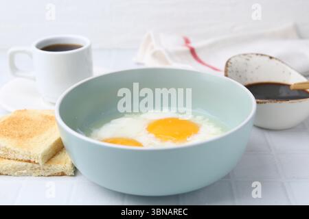 Petit déjeuner asiatique traditionnel avec œufs à moitié durs dans un bol, sauce soja, pain et café sur une table carrelée blanche, gros plan Banque D'Images