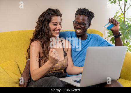 Un jeune couple heureux célèbre un achat ou une réservation en ligne réussi, en tenant une carte de crédit et en utilisant un ordinateur portable. Banque D'Images
