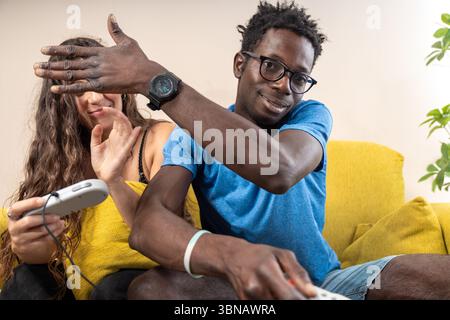 Un homme triche de manière ludique tout en jouant à des jeux vidéo avec sa petite amie, bloquant sa vue et regardant la caméra. Banque D'Images