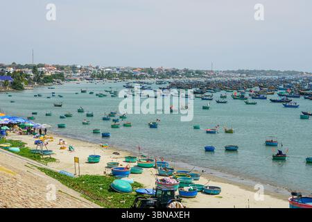 Des centaines de bateaux de pêche dynamiques et de bateaux à panier rond flottent au large de la côte de Mui ne, au Vietnam, près d'un marché côtier animé et d'un paysage urbain Banque D'Images