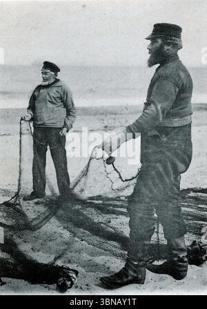 Fisher Life Studies - Looking the nets. Deux personnages se tiennent sur une plage balayée par le vent, chacun tenant un filet de pêche. Vêtus de chandails lourds, de pantalons et de chapeaux à bords, ils font face à la caméra avec une résolution tranquille. La mer s'étend derrière eux, évoquant la vie côtière traditionnelle et les rythmes durables du travail riverain. Publié vers 1904 par Valentine and sons Ltd. D'une carte postale originale. Banque D'Images