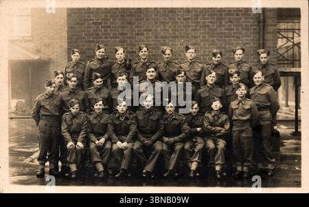 Vingt-sept soldats en uniforme posent en quatre rangées devant un bâtiment en briques, leurs bottes se reflétant sur le sol niché par la pluie. Leurs expressions sont solennelles mais composées. La photographie, probablement au début des années 1900, capture un moment de camaraderie et de discipline au milieu des routines de la vie militaire. D'une carte postale originale, non attribuée et non datée. Banque D'Images