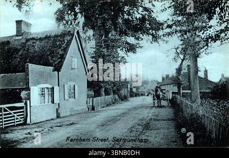 Farnham Street, Saxmundham, capturé dans un moment calme : un chalet au toit de chaume avec une porte blanche se dresse à gauche, tandis que les arbres arquent doucement sur une route non pavée. Une calèche tirée par des chevaux s'arrête en milieu de scène, évoquant une ère pré-automobile de la vie rurale et le charme intemporel du Suffolk. D'après une carte postale originale publiée par H.B. Crisp, non datée mais vers 1910.. Banque D'Images