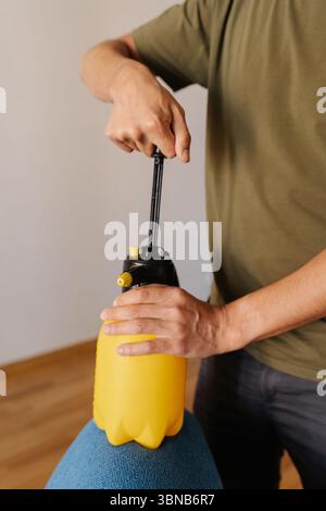 Plan vertical recadré de l'homme pompant la bouteille de pulvérisateur d'eau jaune tout en tenant sur le canapé bleu, se préparant à nettoyer ou désinfecter la sellerie à la maison. Banque D'Images