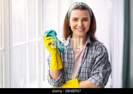 Portrait d'une femme millénaire heureuse avec un chiffon portant des gants en caoutchouc, regardant l'appareil photo et souriant, à l'intérieur Banque D'Images