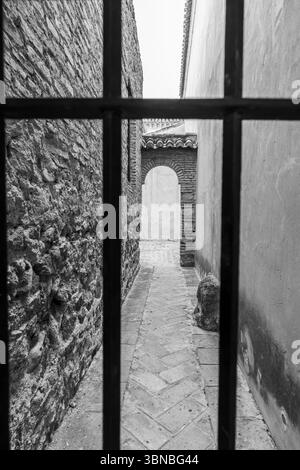 Étroite ruelle en pierre en noir et blanc, vue à travers des barres de fer à l'intérieur de l'Alcazaba de Málaga, évoquant la solitude et l'histoire. Banque D'Images
