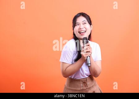 Une femme asiatique joyeuse sourit tout en tenant un micro, regardant loin, prête pour sa performance. isolé sur fond orange Banque D'Images