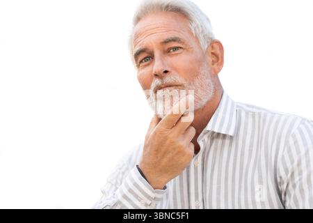 Homme d'affaires mûr avec les cheveux gris et la barbe posant avec une expression réfléchie tout en touchant la barbe et en regardant la caméra sur fond blanc Banque D'Images