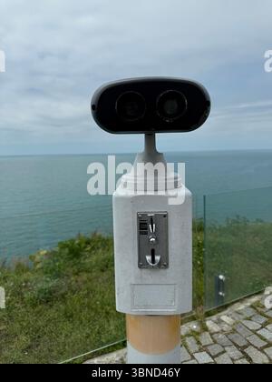 La photo montre une visionneuse binoculaire publique fixe avec une fente pour pièces de monnaie, positionnée sur une plate-forme en bord de mer. Le fond comprend la mer et un ciel nuageux, Banque D'Images