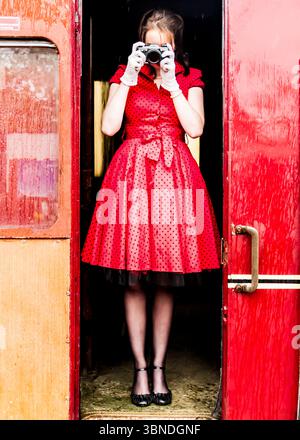 Une femme dans une robe à pois rouges et des gants blancs se tient devant la porte d'un wagon rouge, tenant un appareil photo sur son visage. Banque D'Images