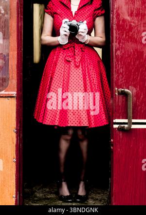 Une femme dans une robe à pois rouges et des gants blancs tient un appareil photo devant elle, debout dans une porte. Banque D'Images