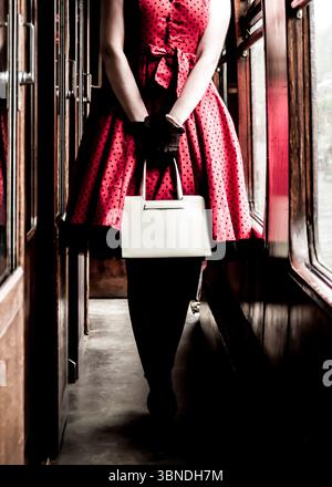 Une femme dans une robe à pois rouges et des gants noirs se tient dans un wagon, tenant un sac à main blanc. Banque D'Images