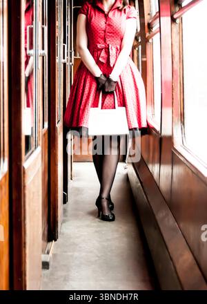 Une femme dans une robe à pois rouges, des gants noirs et des talons se tient debout dans un wagon de train vintage, tenant un sac à main blanc. Banque D'Images