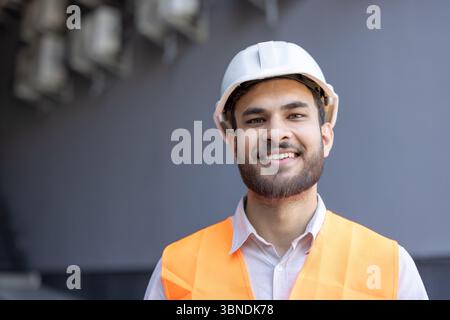 Un travailleur de la construction souriant porte un casque blanc et un gilet de sécurité orange, regardant directement la caméra. Banque D'Images